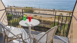 Ein Balkon mit zwei Korbstühlen und einem kleinen runden Tisch, mit Blick auf einen Strand mit Menschen und Palmen.