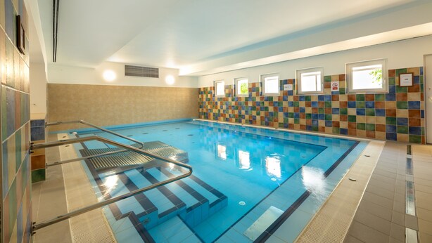 An indoor swimming pool with a tiled wall, a metal ladder, and a window. - Hotel Szárcsa (Szekesfehervar)