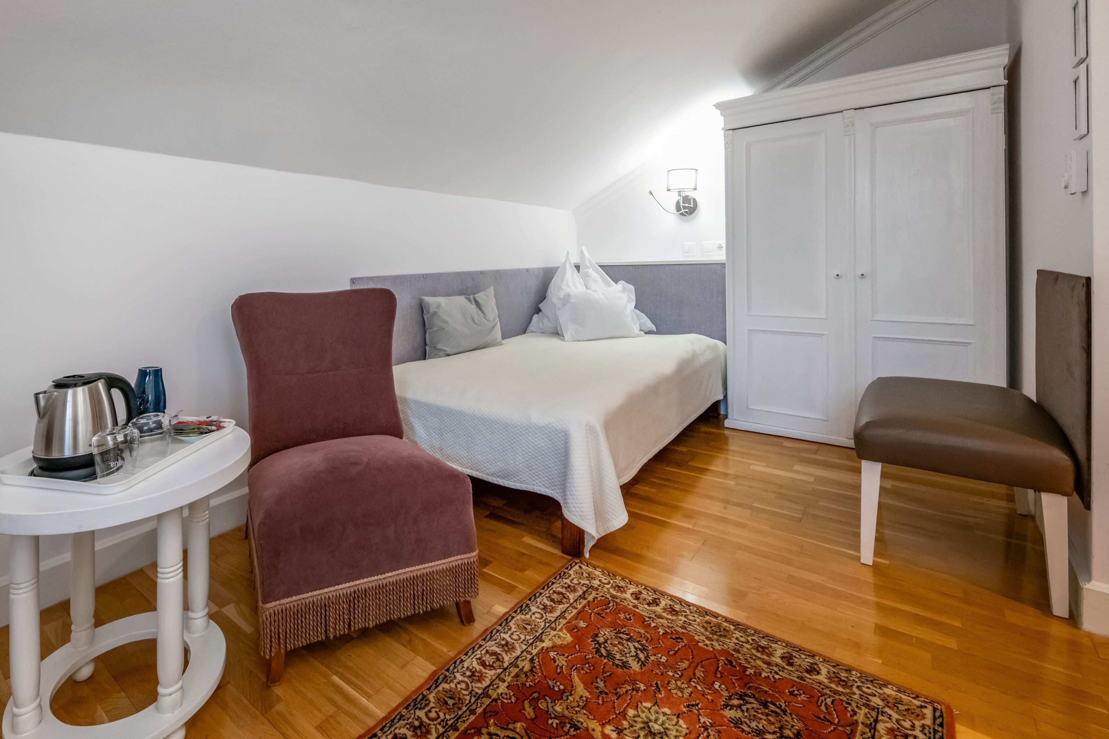 A bedroom with a bed, a chair, a small table with a kettle, and a rug.