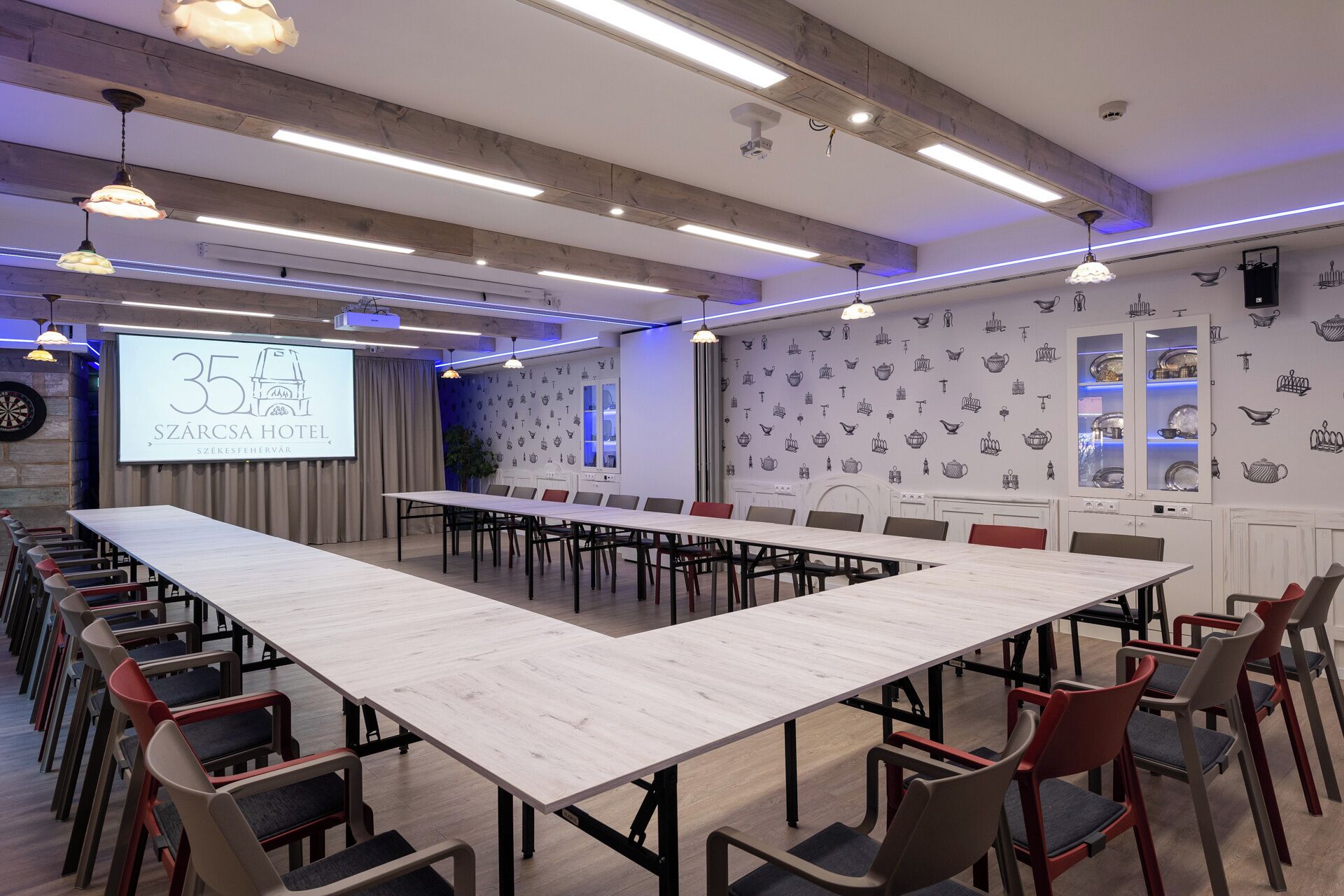 A conference room with a long table, multiple chairs, a projector screen, and a wall with kitchenware illustrations.