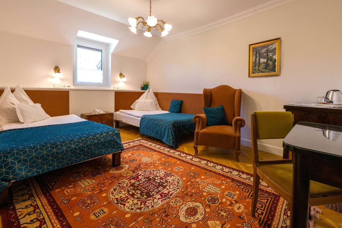 Ein Hotelzimmer mit zwei Betten, einem Sessel, einem Tisch, einem Schreibtisch, einem Gemälde, einer Chandelier und einem Fenster.