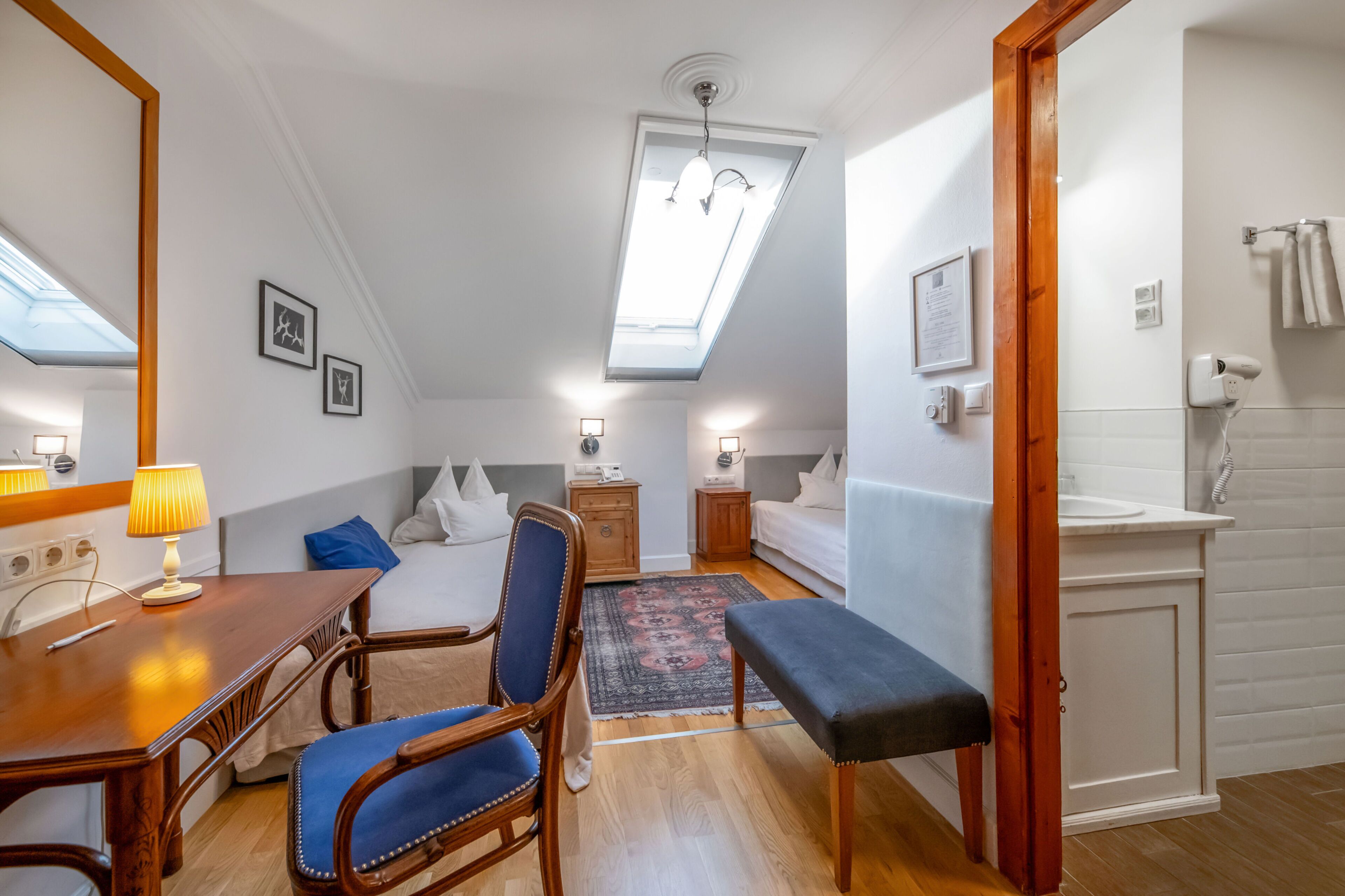 A bedroom with a skylight, a desk with a lamp, a chair, a bedside table, a bed, a bedside table, a rug, a bench, a wardrobe, and a bathroom with a towel rack.