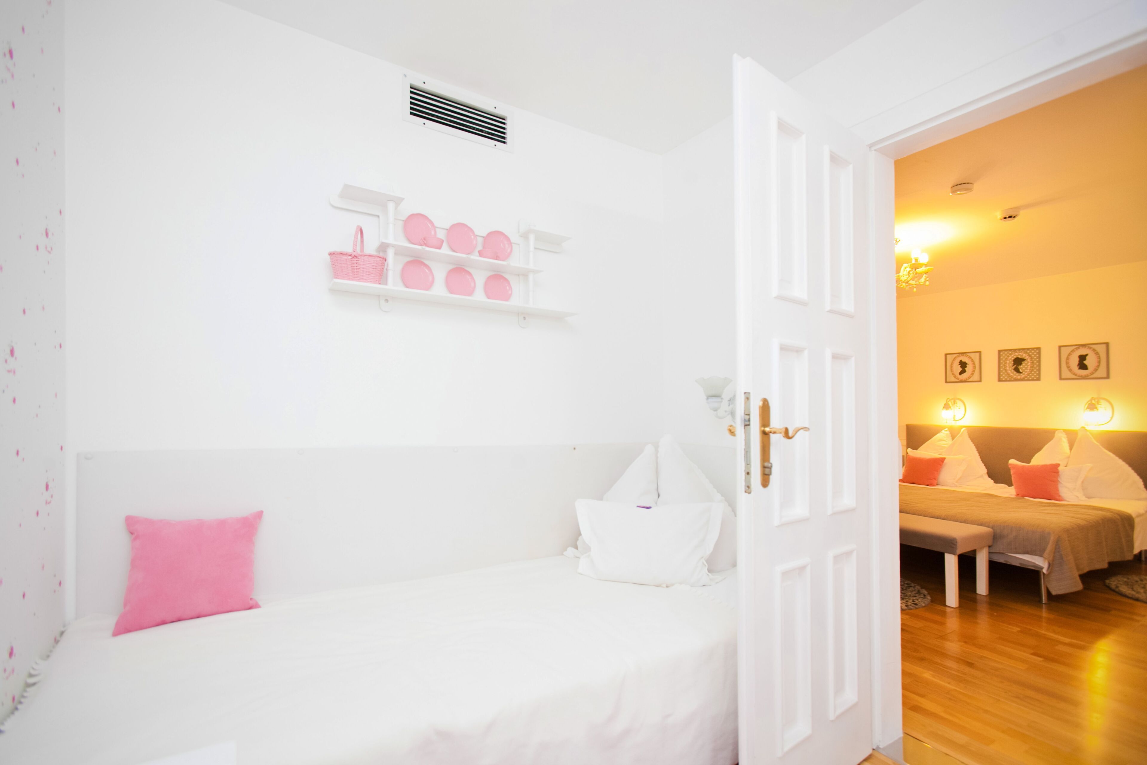 Una habitación con una cama blanca, un banco, una puerta que da a otra habitación y un estante en la pared con objetos rosados.