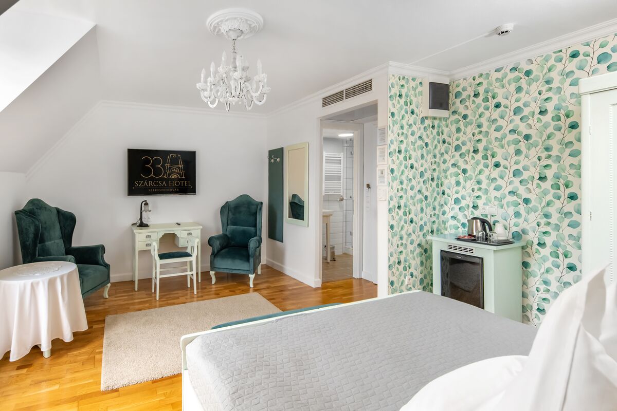 A bedroom with a bed, a desk, two chairs, a chandelier, a small table, and a wall-mounted TV.