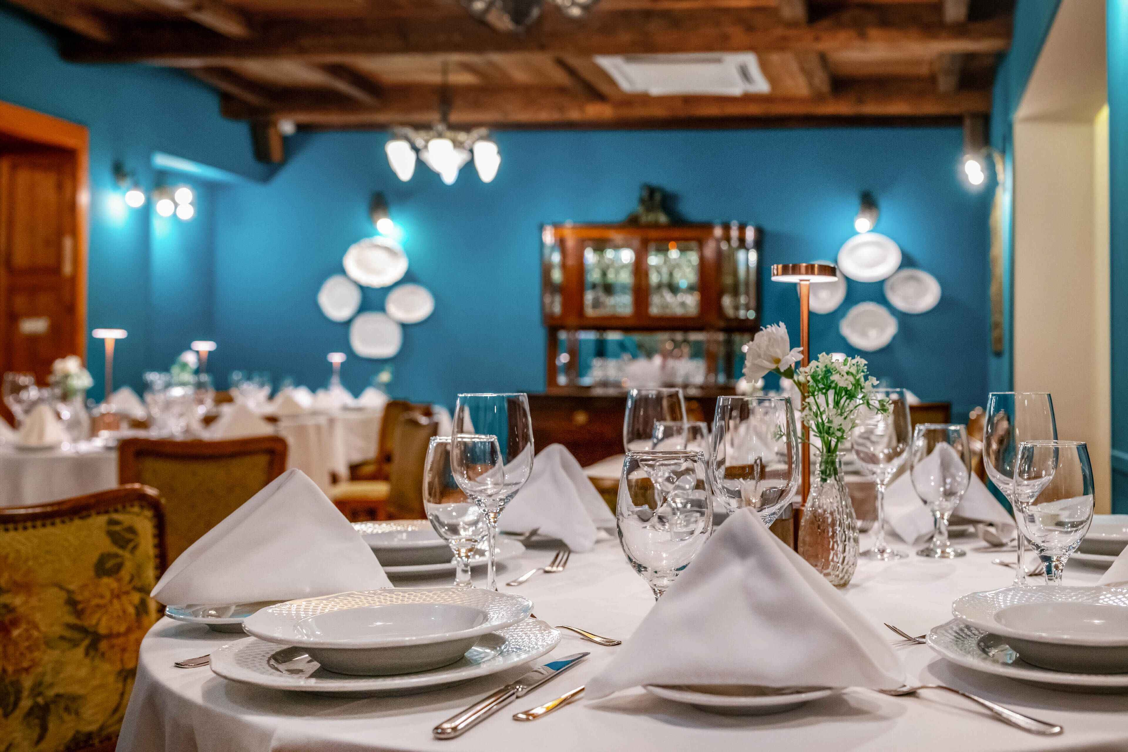 A dining table set with plates, glasses, and cutlery, with a blue wall and wooden ceiling in the background.