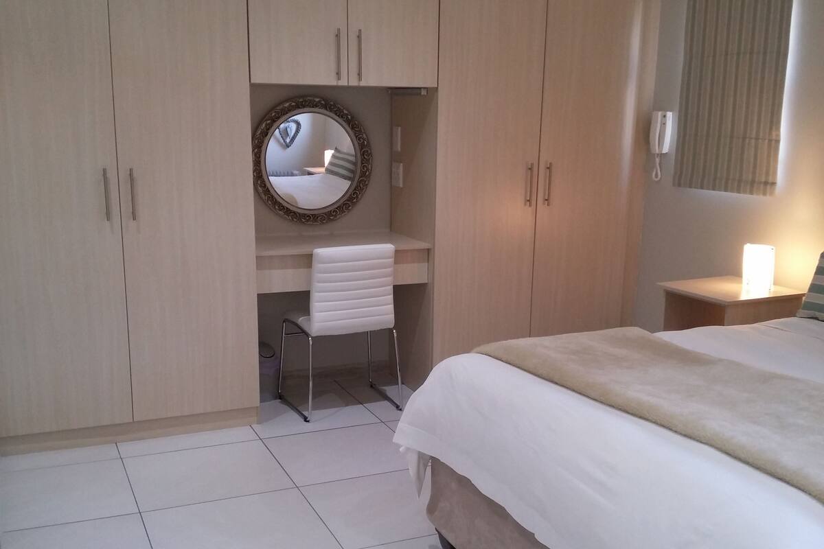 A bedroom with a bed, a desk, a mirror, and a wardrobe.
