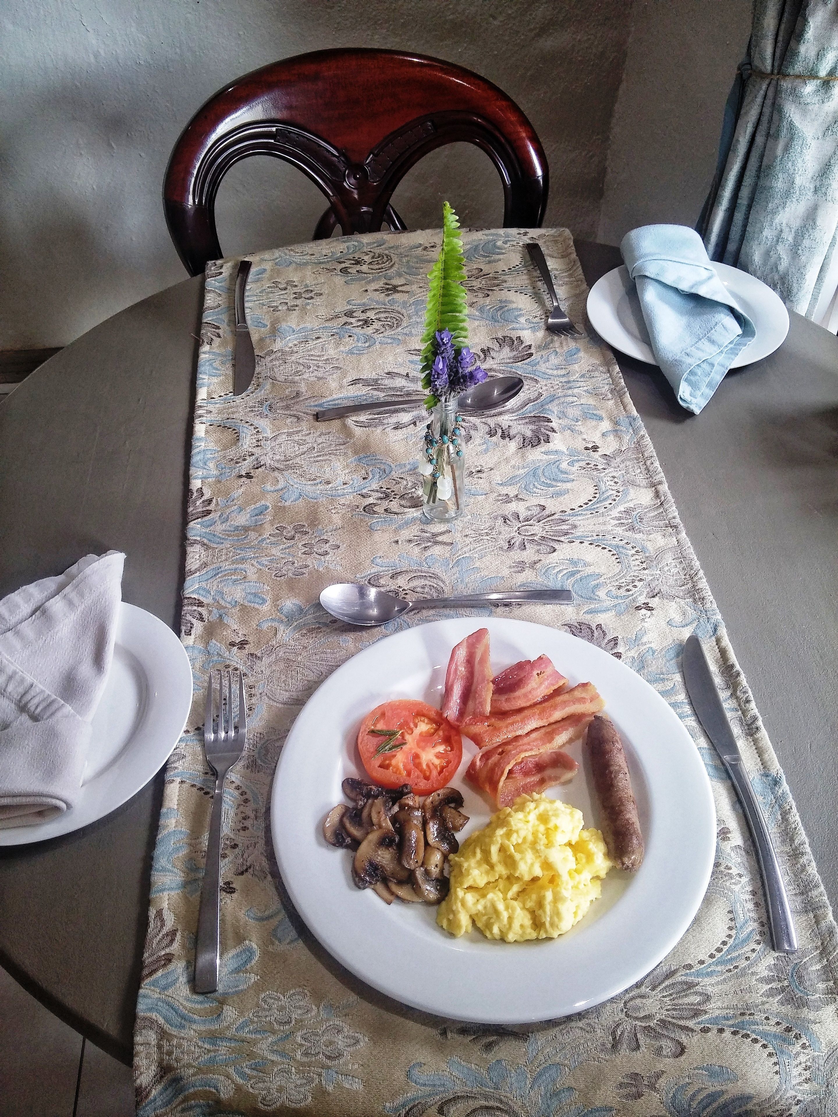 A breakfast table with a plate of eggs, bacon, mushrooms, and a tomato.