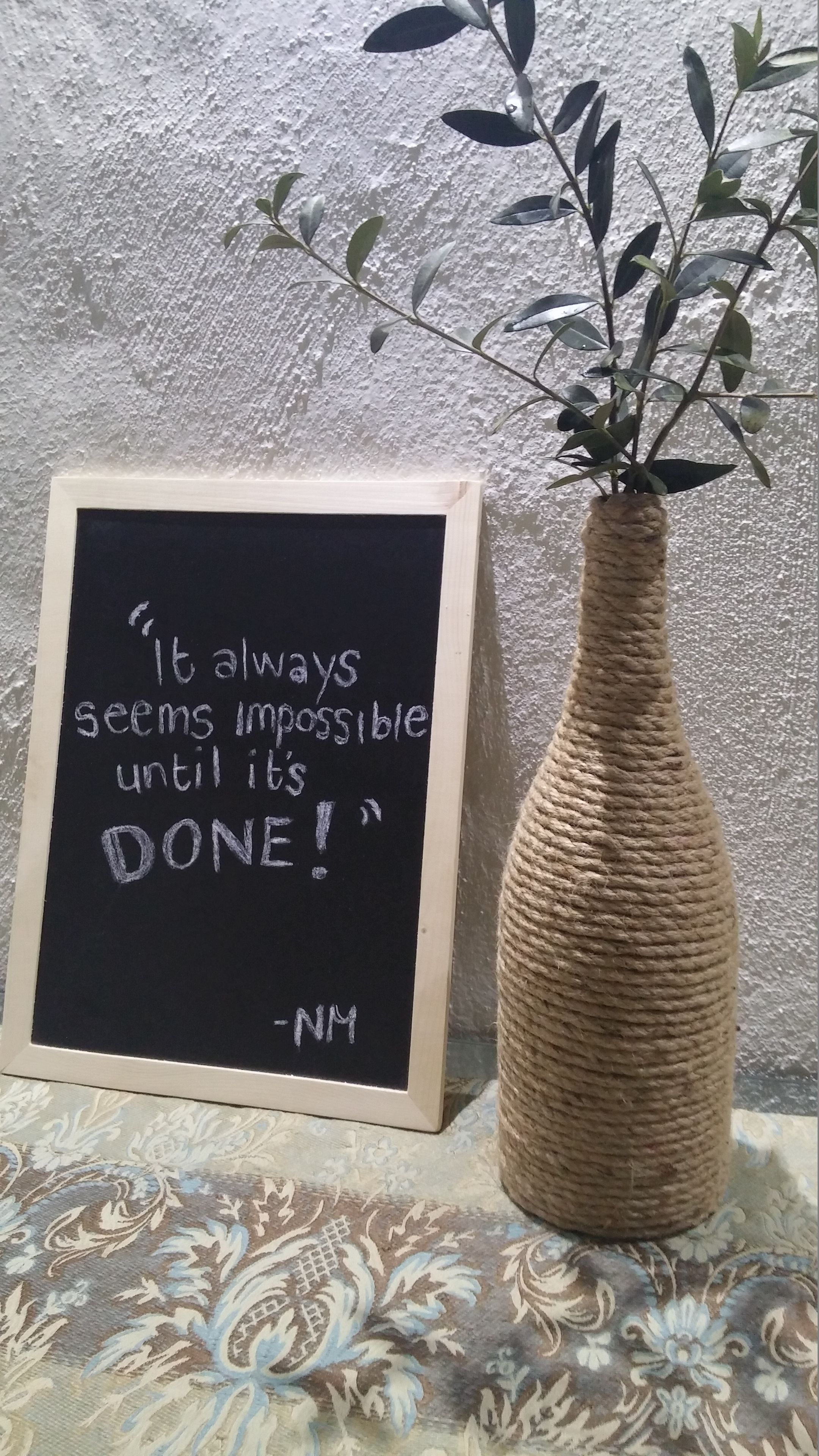 Ein gerahmter Spruch an einer Schiefertafel neben einer gewebten Vase mit einer Pflanze.