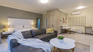 Une chambre moderne avec un grand lit, un canapé gris, une petite table ronde avec une plante et un coin cuisine.