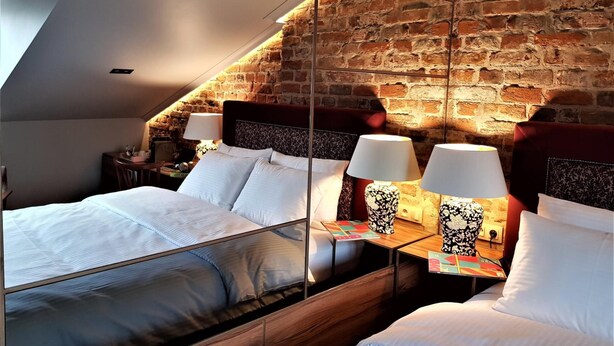 A bedroom with a sloped ceiling, a brick wall, and a mirrored headboard reflecting the room. There are two bedside tables with lamps and a bed with white bedding. - SleepWell Apartments Nowy Świat (Warsaw)