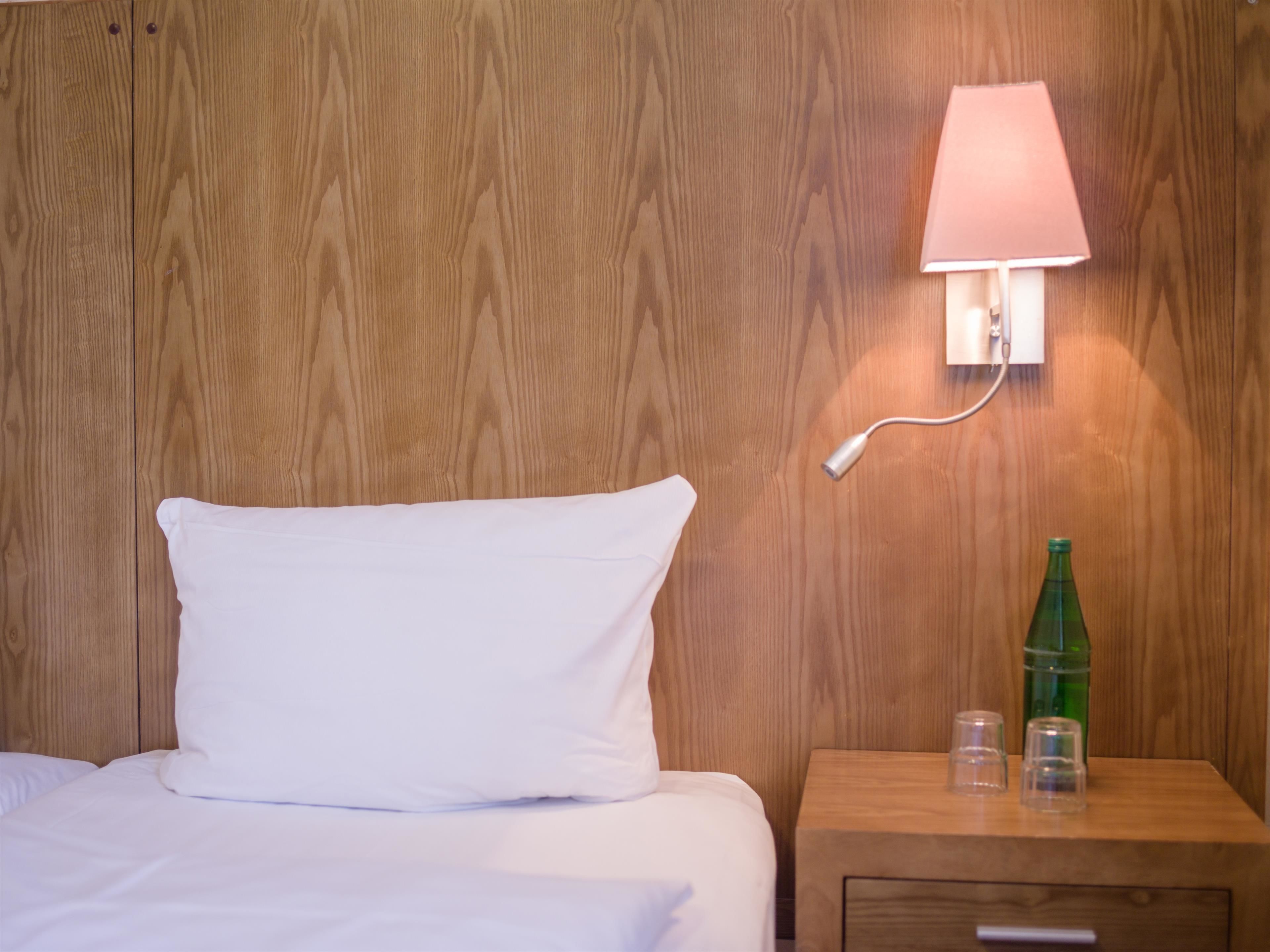 A wooden wall with a white pillow on a bed, a bedside table with a green bottle and two glasses, and a wall-mounted lamp.