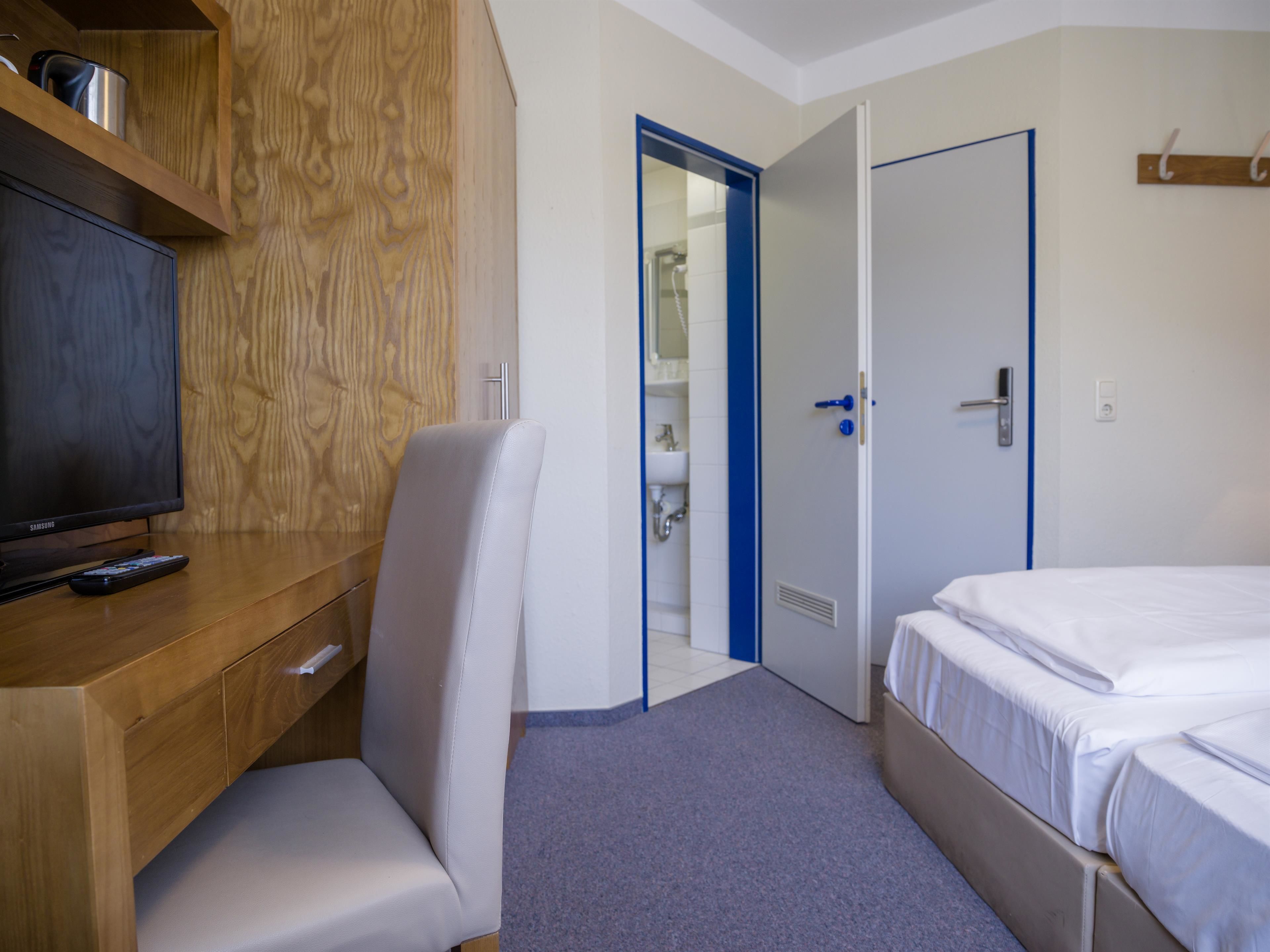 Habitación de hotel con un escritorio de madera, una silla, una cama y un baño visible a través de una puerta abierta.