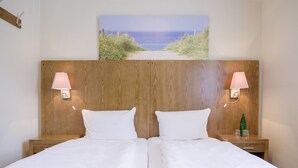 Cama doble con ropa de cama blanca, cabecera de madera, dos lámparas de noche y un cuadro con una escena de playa sobre la cama.