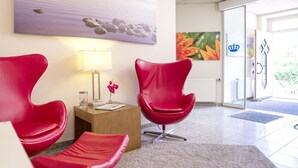 Un área de recepción con dos sillones rojos, una mesita con una lámpara, un jarrón con flores y un cuadro en la pared.