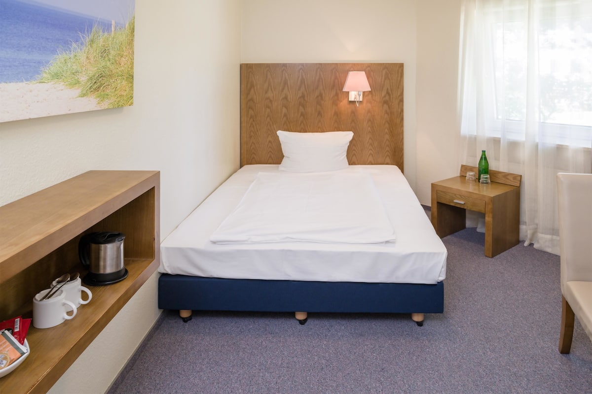 Una cama bien hecha con cabecera de madera, una mesita de noche con lámpara, un estante con tetera y tazas, y un cuadro de una playa en la pared.