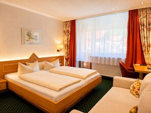 A hotel room with a double bed, a small table, a chair, a desk, and a window with curtains. - Hotel Arnika (Oberammergau)