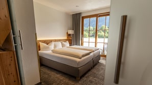 A hotel room with a large bed, a wooden headboard, a window with a view of trees, and a door with a wooden frame. - Hotel Arnika (Oberammergau)