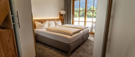 Een hotelkamer met een groot bed, een houten hoofdbord, een raam met uitzicht op bomen en een deur met houten omlijsting.
