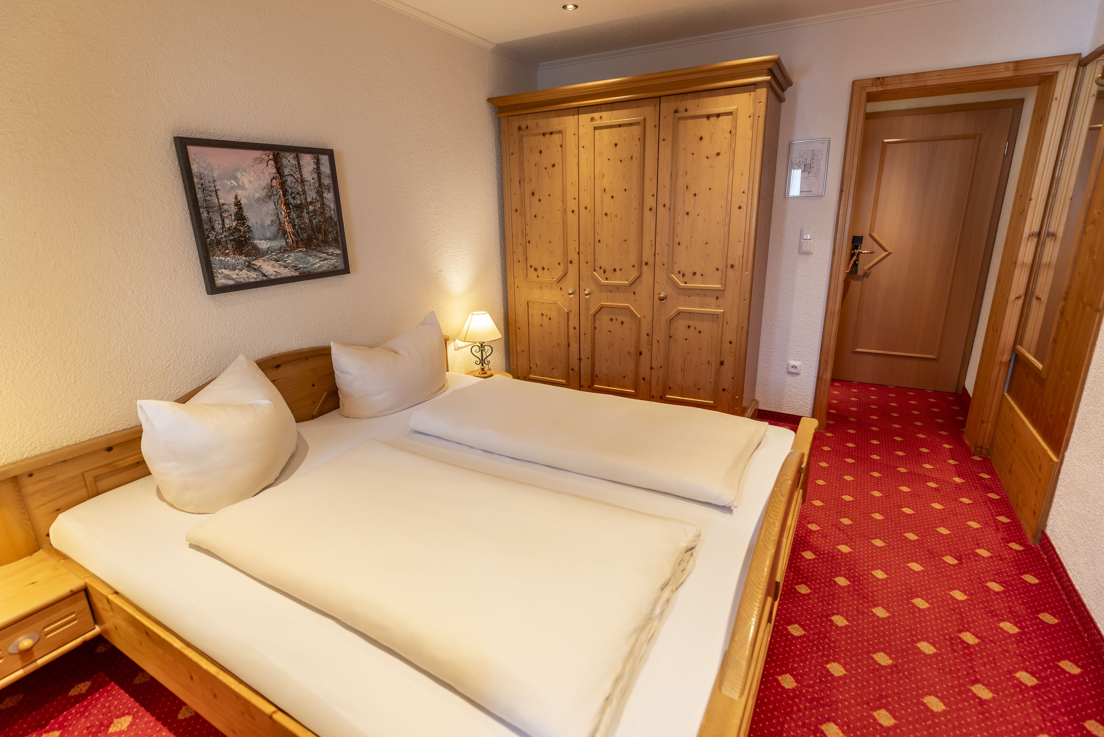 A double bed with white linens, a wooden wardrobe, a painting of a snowy landscape, and a red patterned carpet.