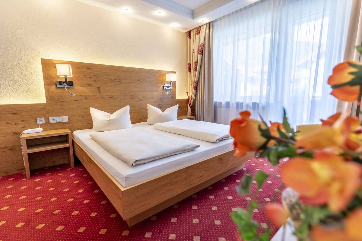 A hotel room with a wooden headboard, two beds with white linens, a nightstand with a lamp, and a window with sheer curtains.