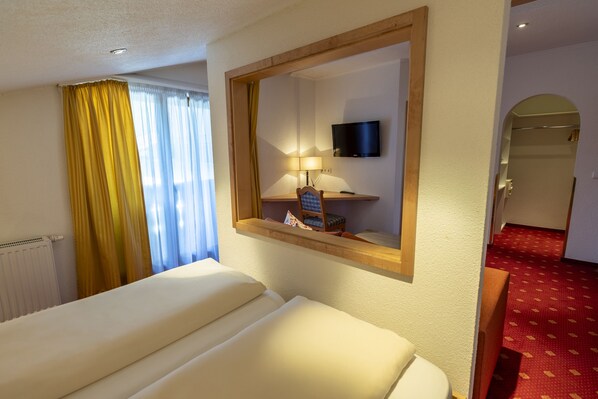 Una habitación de hotel con cama, televisión montada en la pared, lámpara y ventana con cortinas.