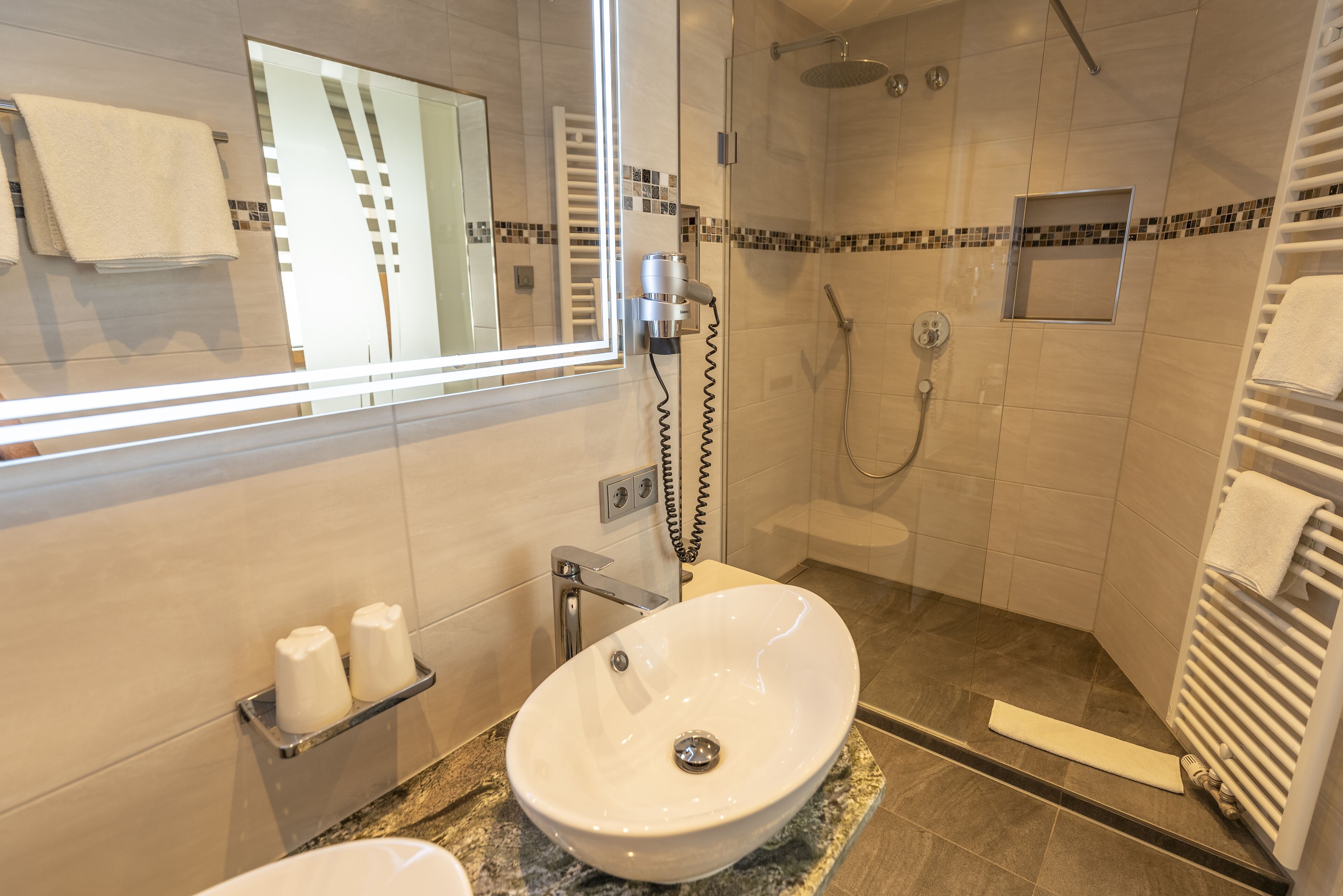 A modern bathroom with a large mirror, a white sink, a shower area, and a towel rack.