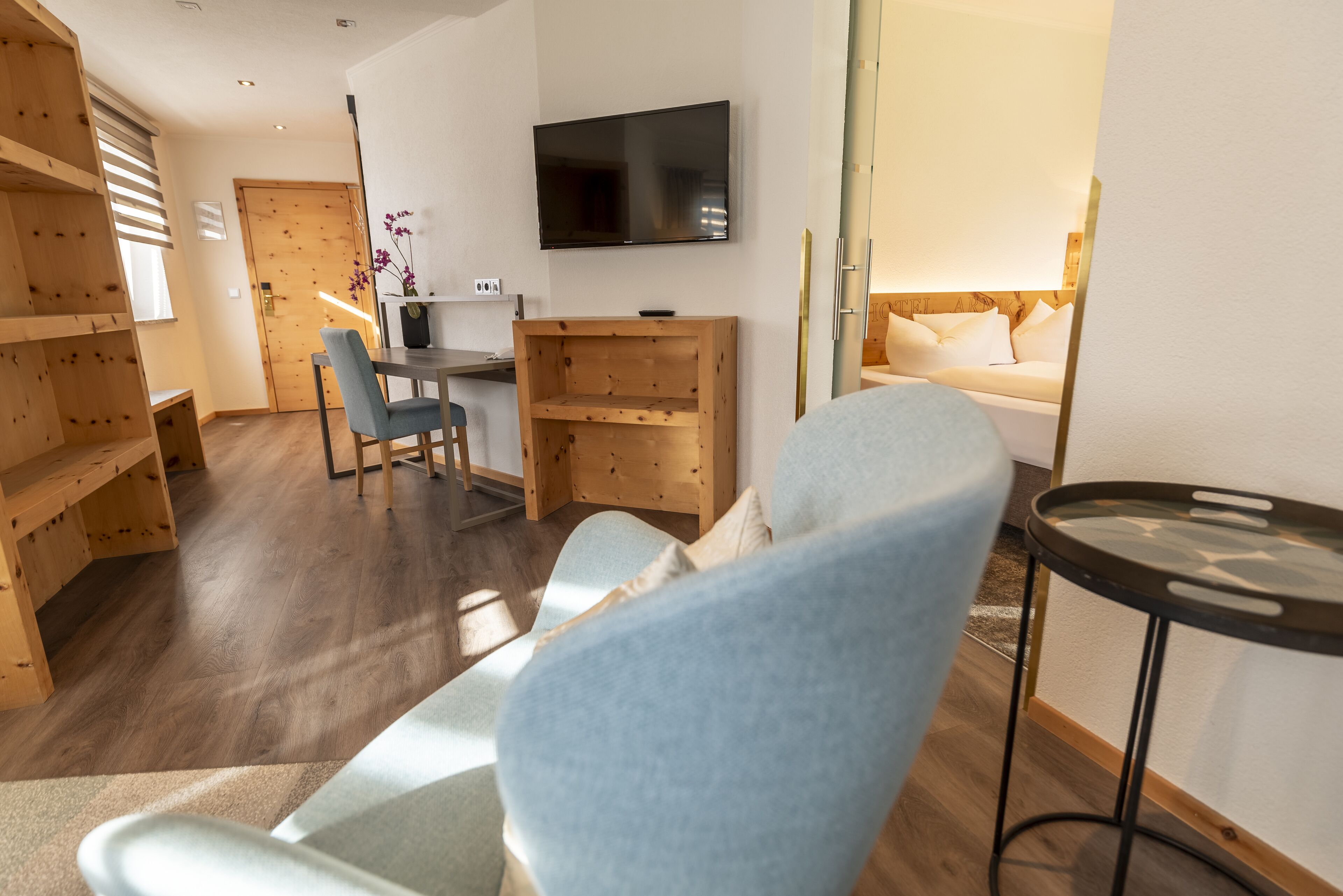 A modern hotel room with a wooden floor, a flat-screen TV mounted on the wall, a dining area with a table and chairs, and a bed with a headboard.