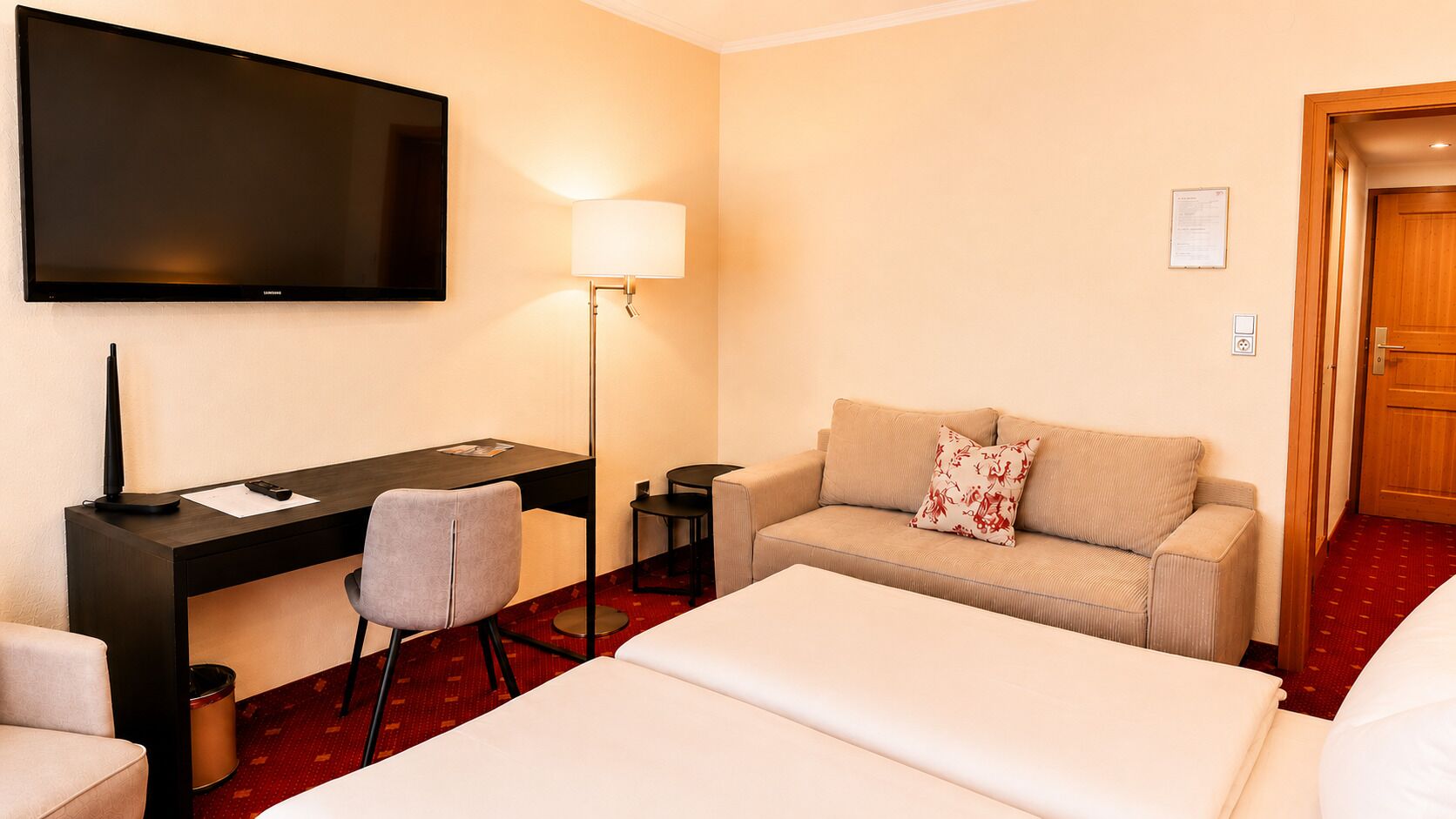 Habitación de hotel con cama, sofá, escritorio con silla, televisor montado en la pared y lámpara.