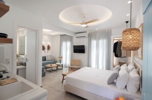 A modern hotel room with a large bed, a flat-screen TV, a ceiling fan, and a dining area with a table and chairs. - Agia Anna Living (Naxos)