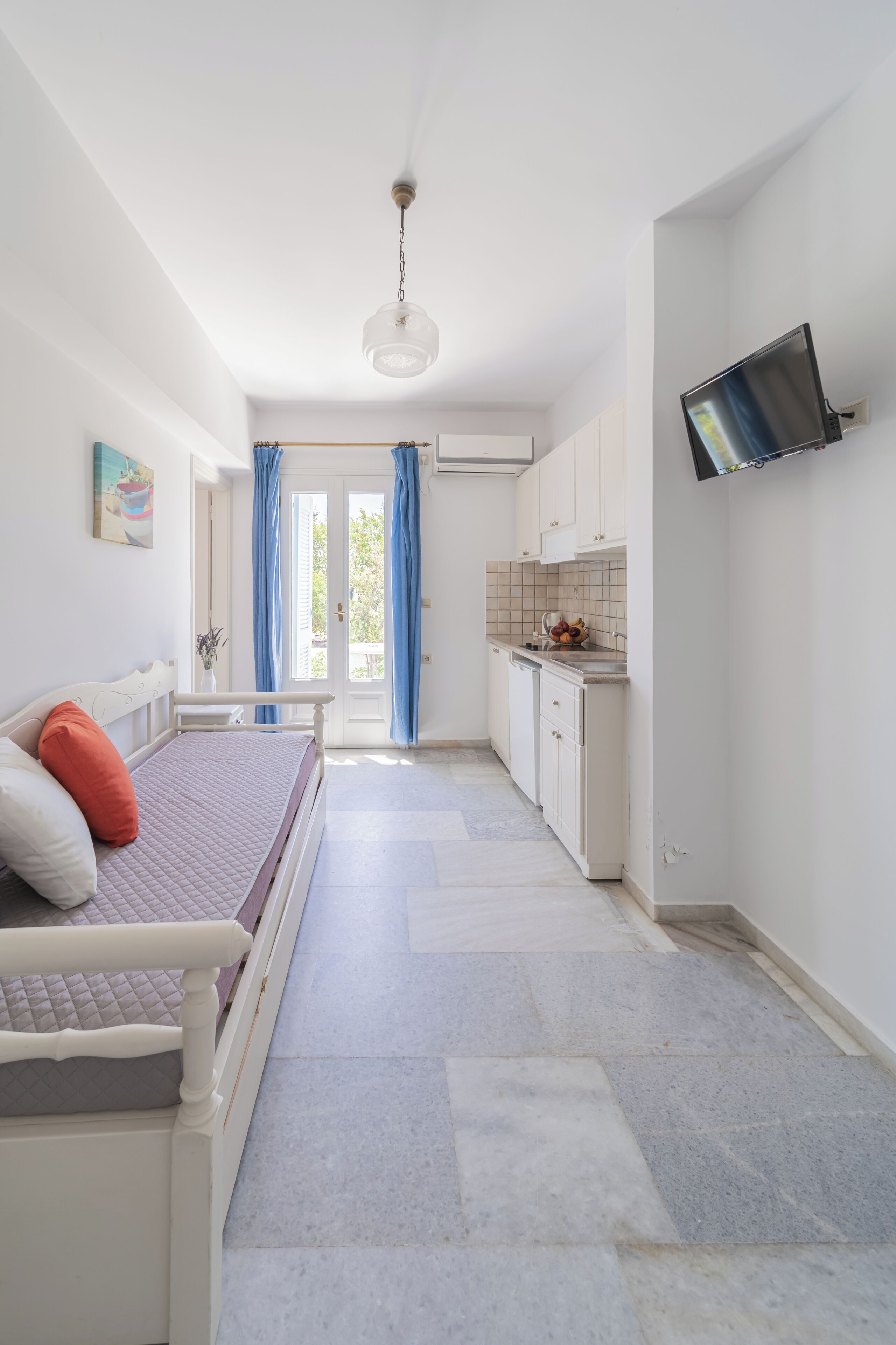 A bedroom with a bed, a TV mounted on the wall, and a kitchen area with cabinets and a sink.
