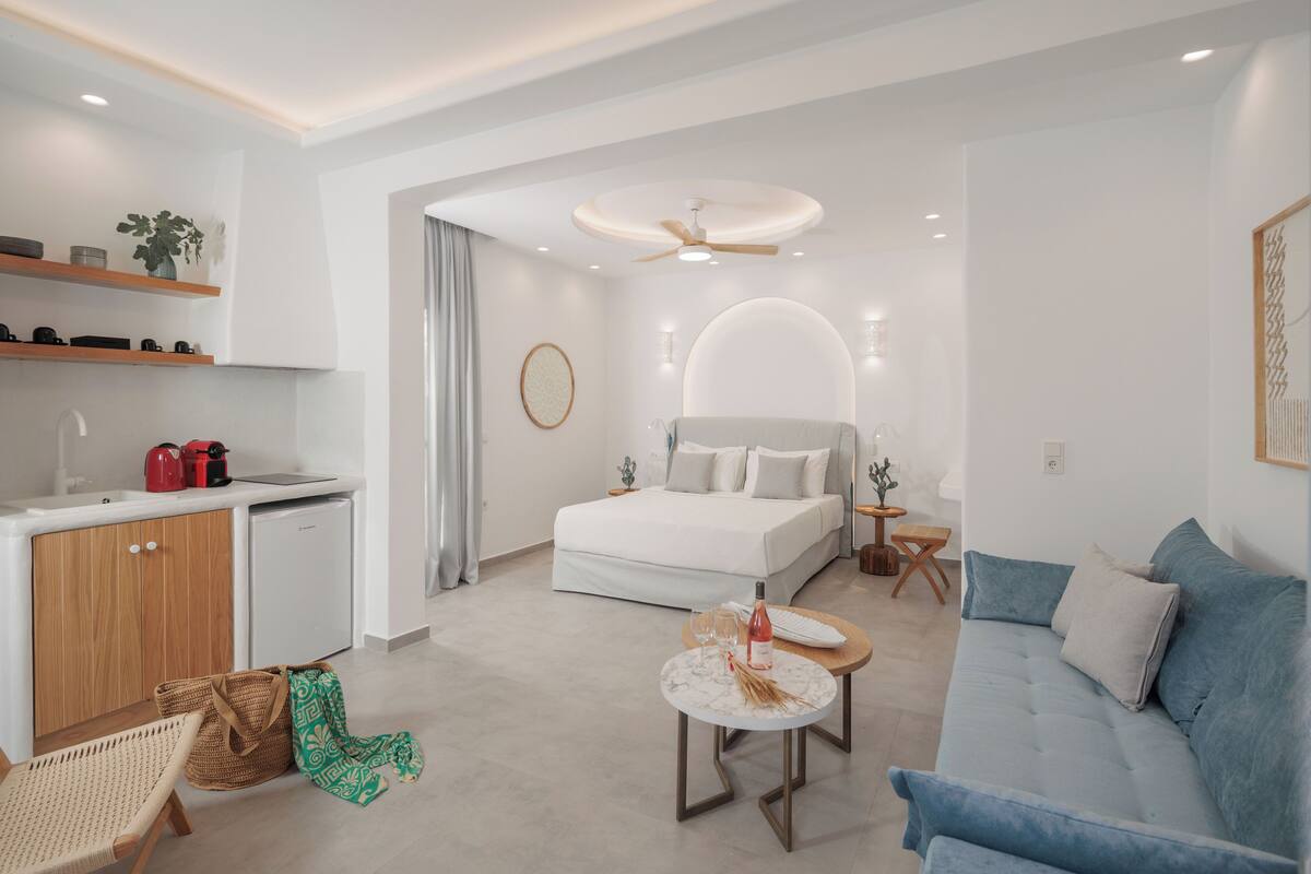 A modern hotel room with a bed, a sofa, a small kitchen area, and a dining table.