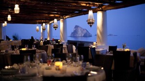 Un restaurante con mesas dispuestas para comer, con vista a una masa de agua y una isla rocosa en la distancia.