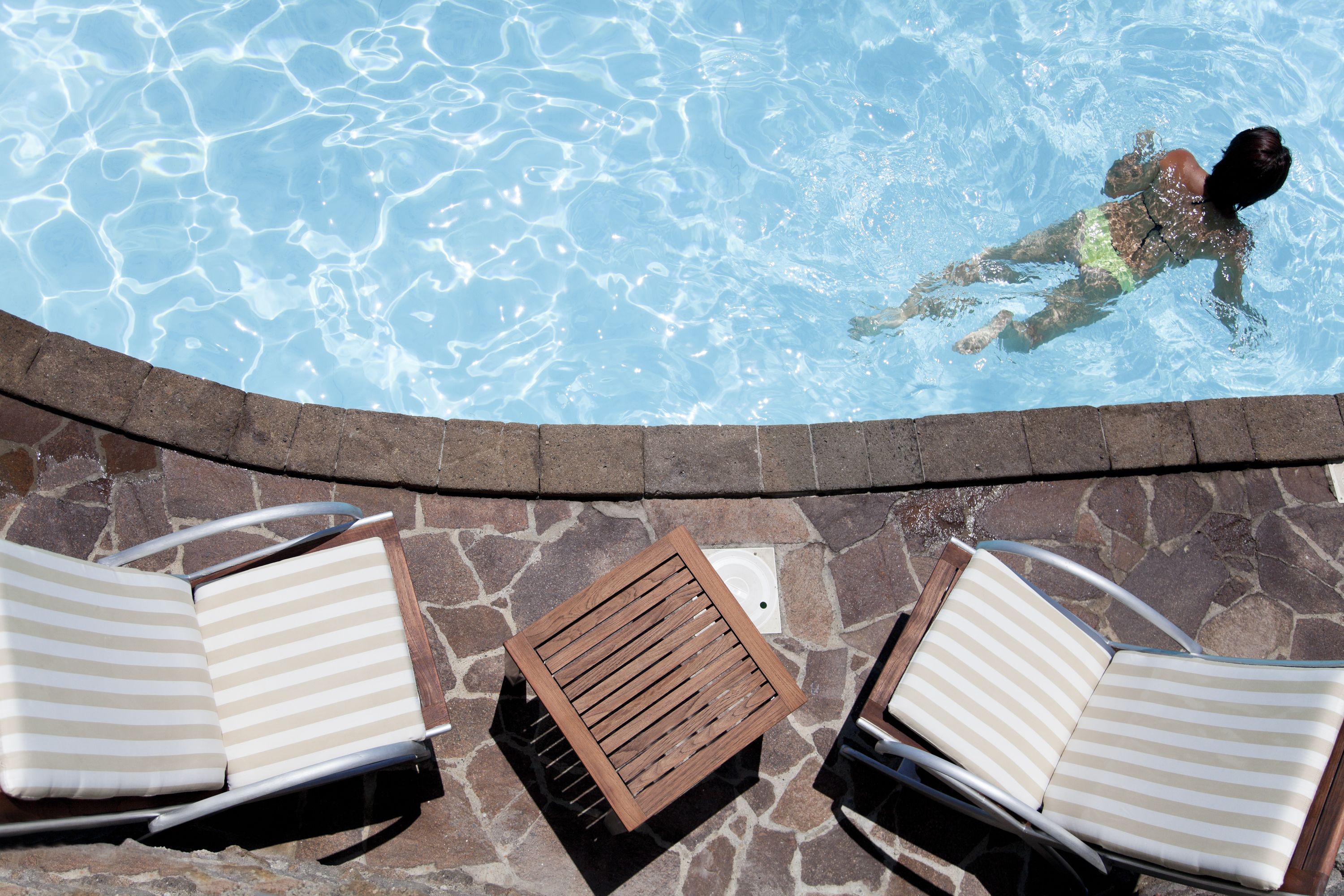 A swimming pool with a person swimming, two sun loungers, and a stone-tiled deck.