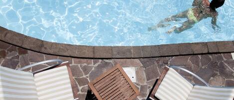 A swimming pool with a person swimming, two sun loungers, and a stone-tiled deck.