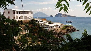 Un edificio costero con varios balcones y una zona de piscina, con vistas al mar y una montaña al fondo.