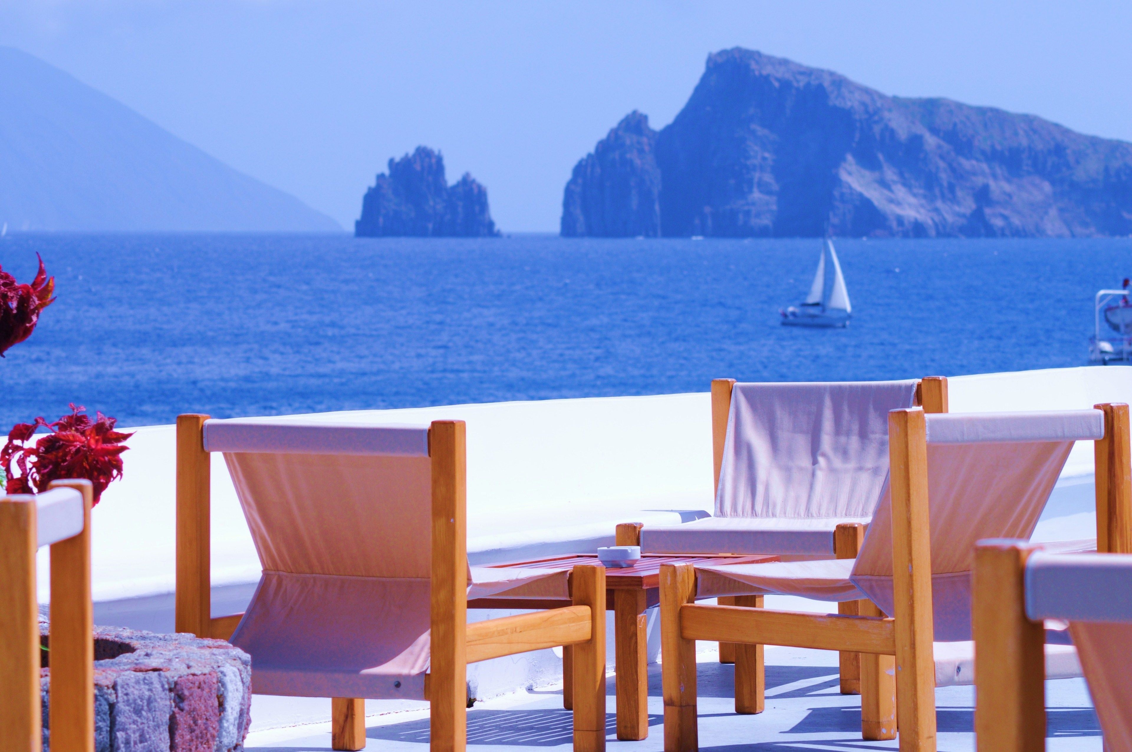 Une terrasse en bord de mer, avec des chaises et des tables en bois, d’où l’on peut admirer la mer et un voilier.