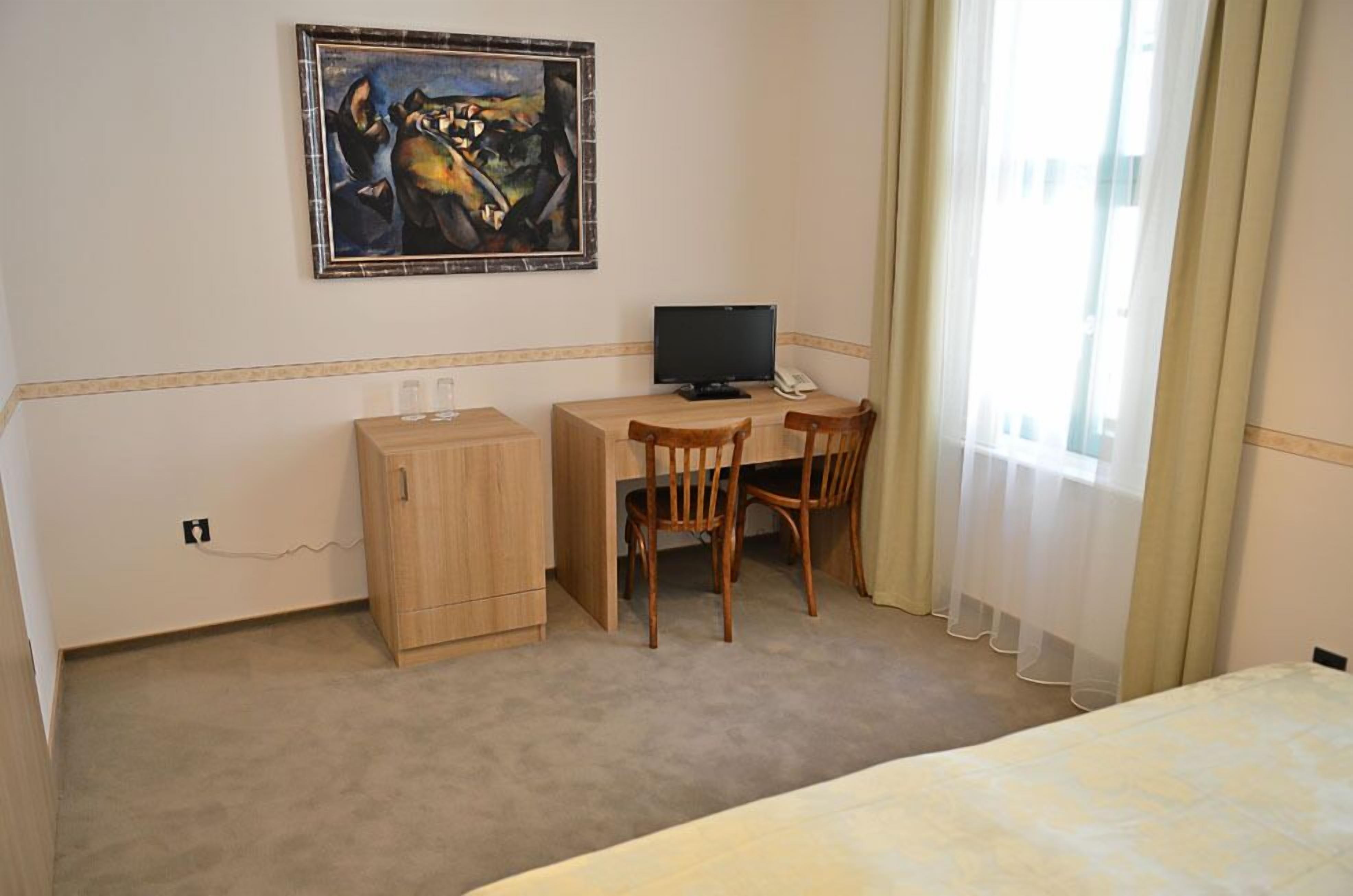 Une chambre d’hôtel avec un lit, un bureau avec un ordinateur, une chaise, un petit meuble et un tableau au mur.