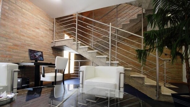 A modern interior with a glass coffee table, white sofas, a TV, and a staircase against a brick wall. - Hotel Arkadia (Pecs)