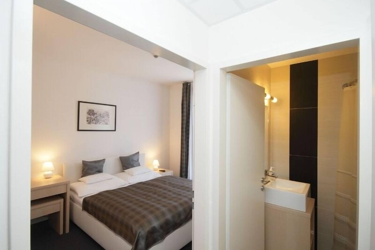 Habitación de hotel con cama, mesitas de noche, un cuadro en la pared y un baño con lavabo y espejo.