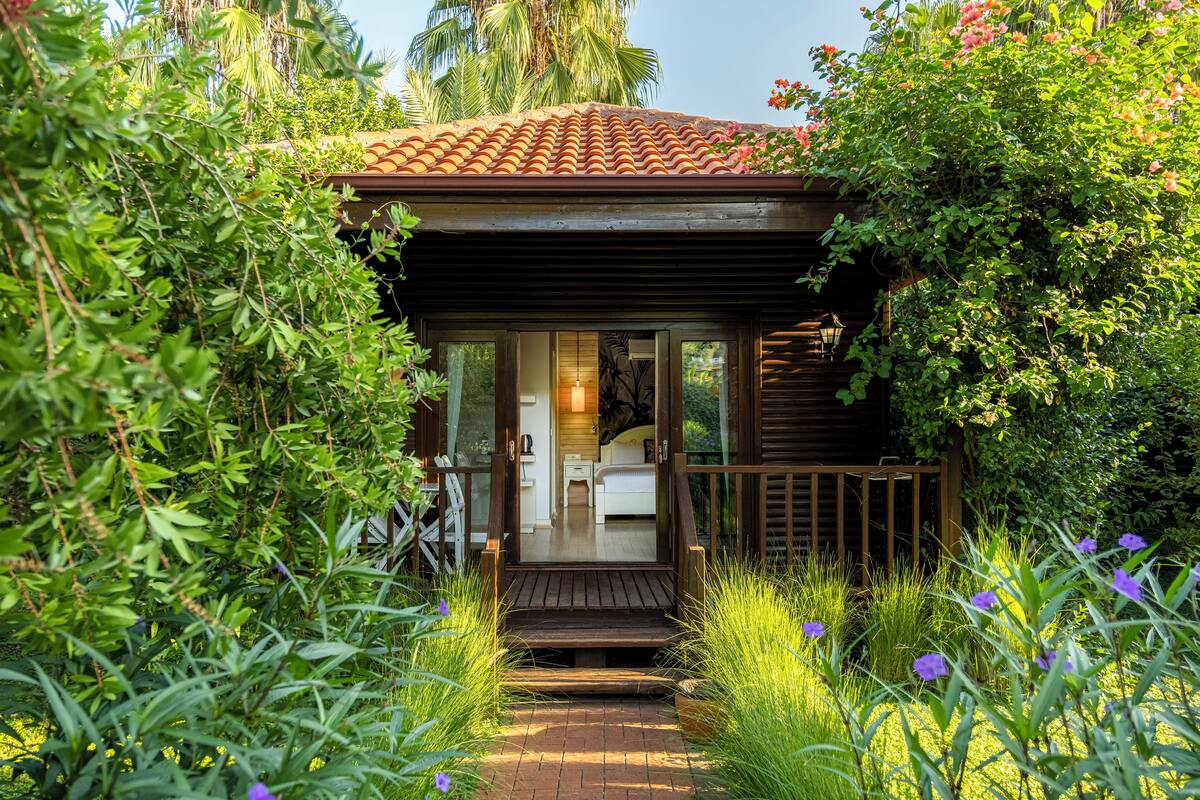 A small wooden cabin with a tiled roof, surrounded by lush greenery and flowering plants.