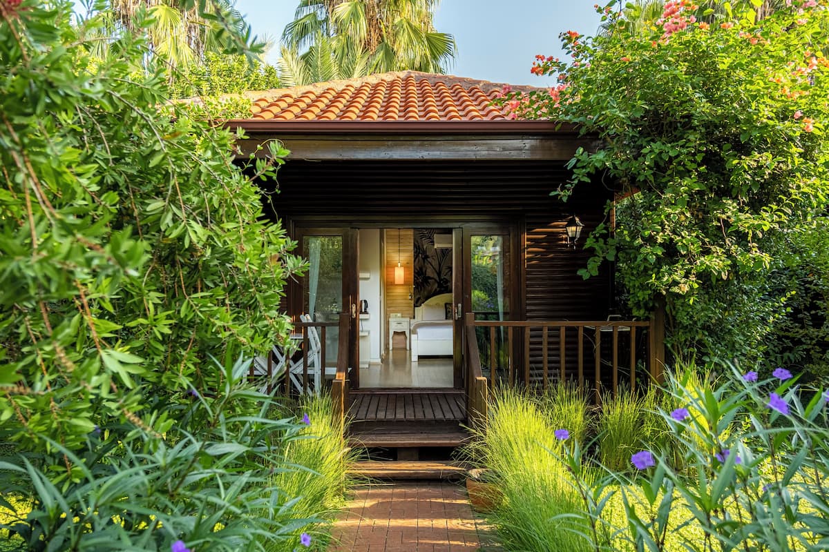 Une petite cabane en bois, surmontée d’un toit de tuiles, entourée d’une végétation luxuriante et de plantes en fleurs.