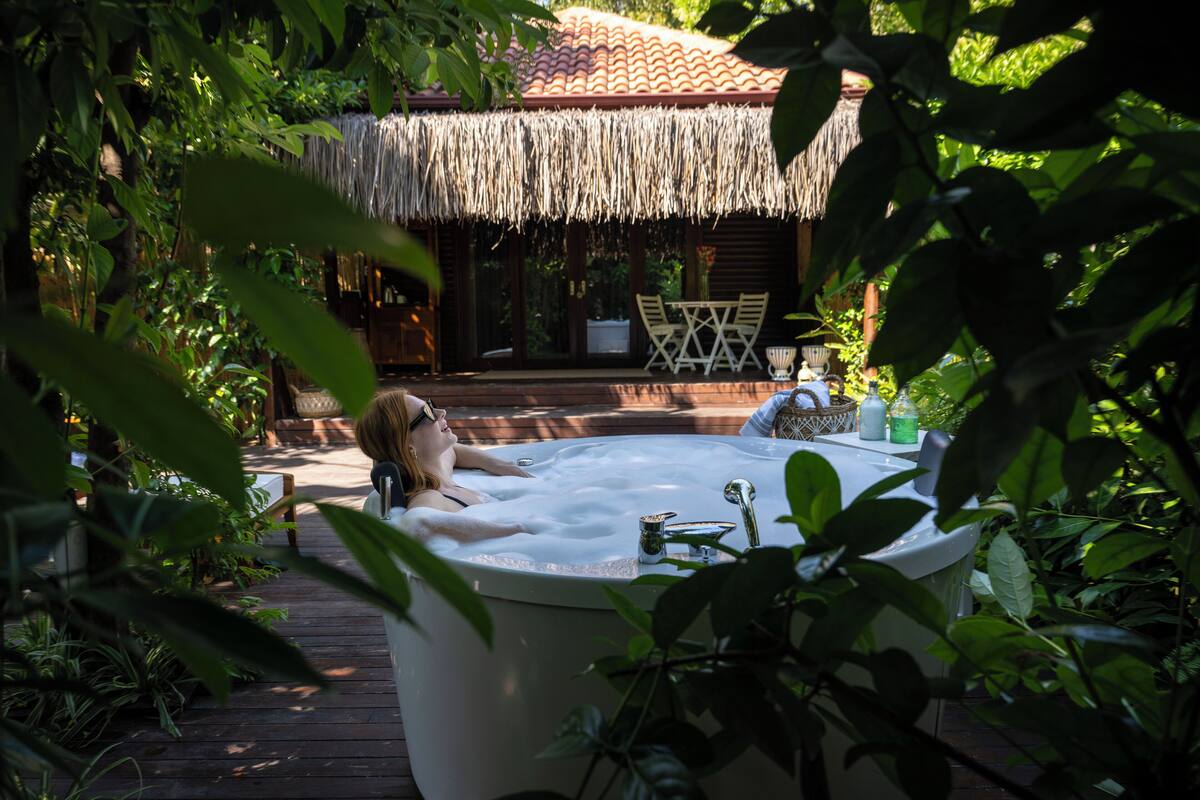 A person relaxing in a hot tub surrounded by lush greenery and a thatched-roof structure.
