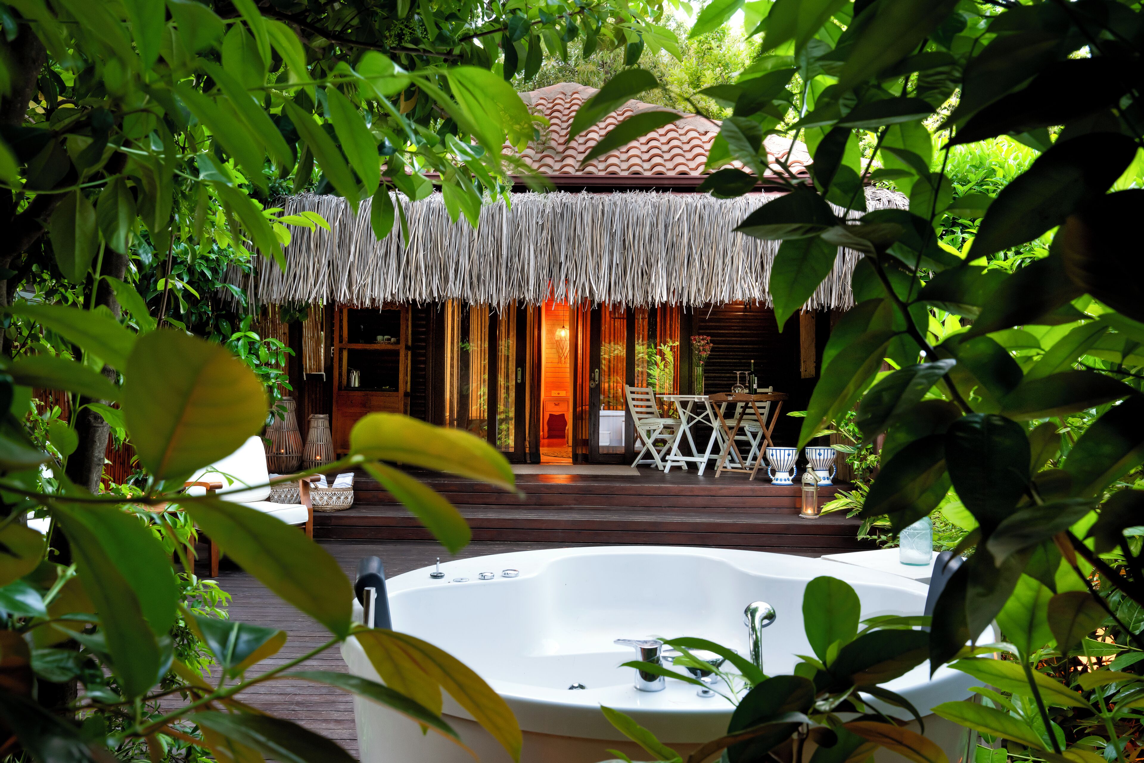 A thatched-roof structure with a wooden deck, a hot tub, and outdoor furniture.