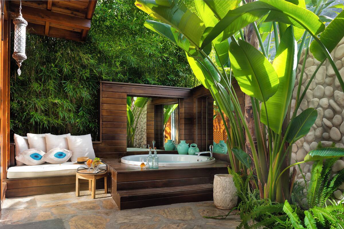 A spa area with a wooden bench, a stone floor, a bathtub, and lush greenery.