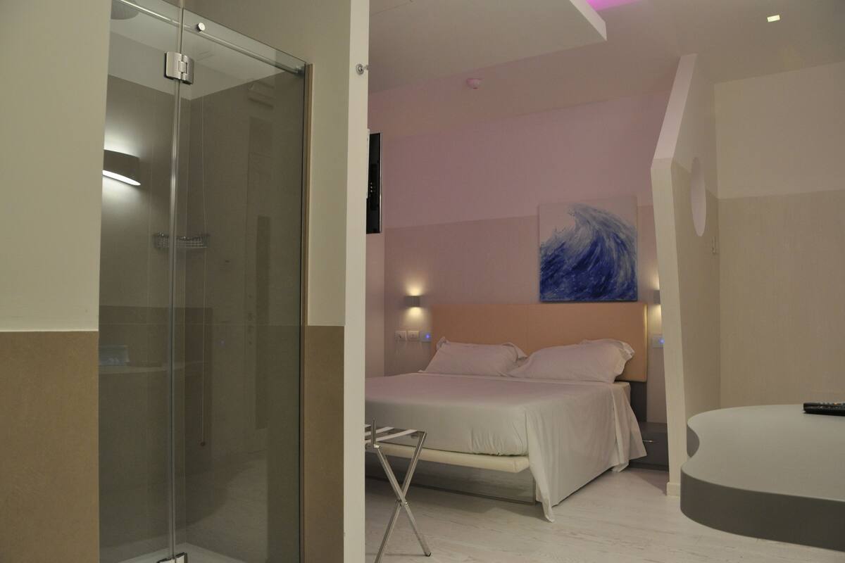 Ein modernes Hotelzimmer mit einer gläsernen Dusche, einem Bett mit weißen Leinen, einem an der Wand montierten Fernseher und einem Gemälde.
