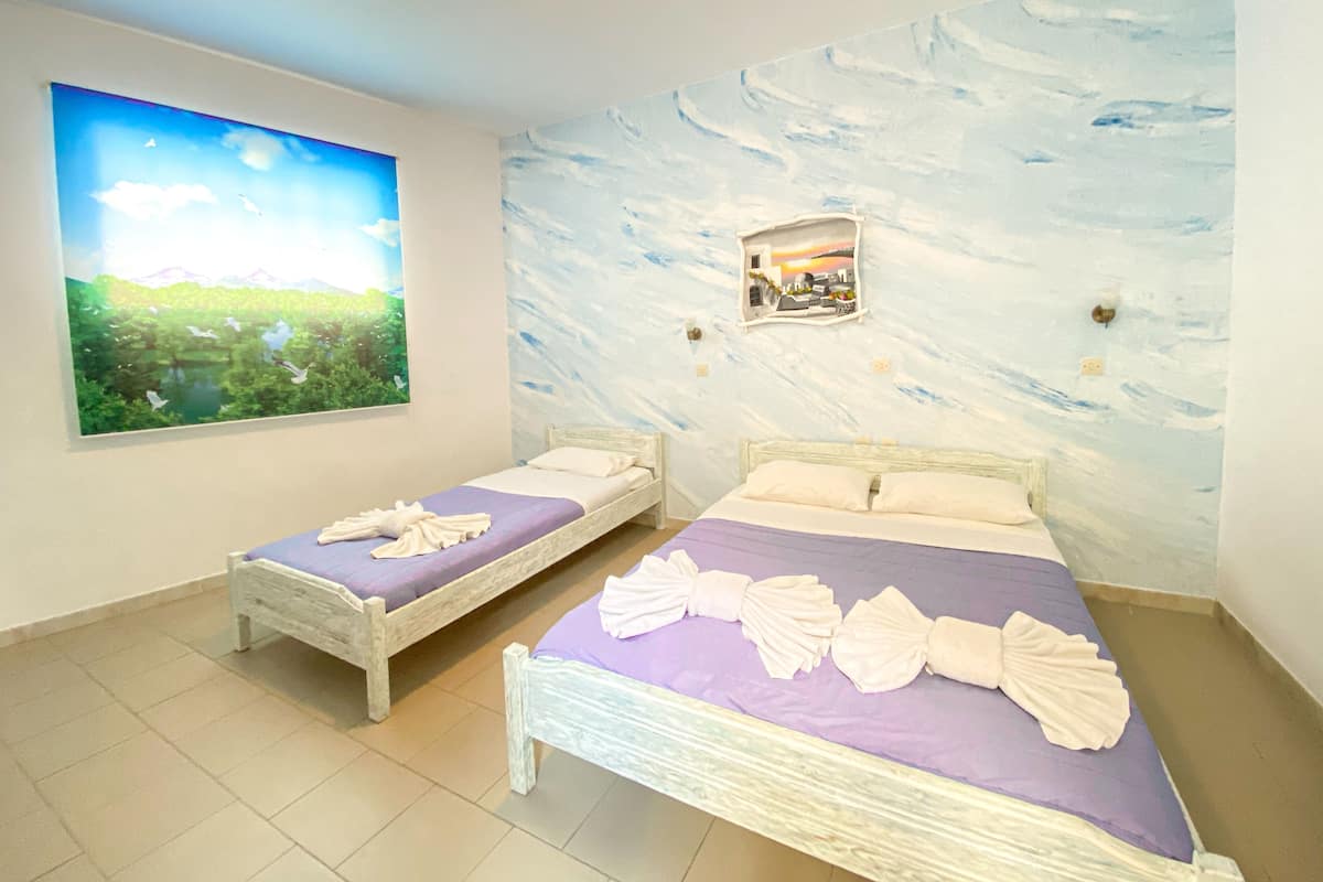 Une chambre avec deux lits, chacun recouvert d’une courtepointe violette et agrémenté d’oreillers blancs. Un tableau représentant un paysage est accroché au mur, à gauche des lits.