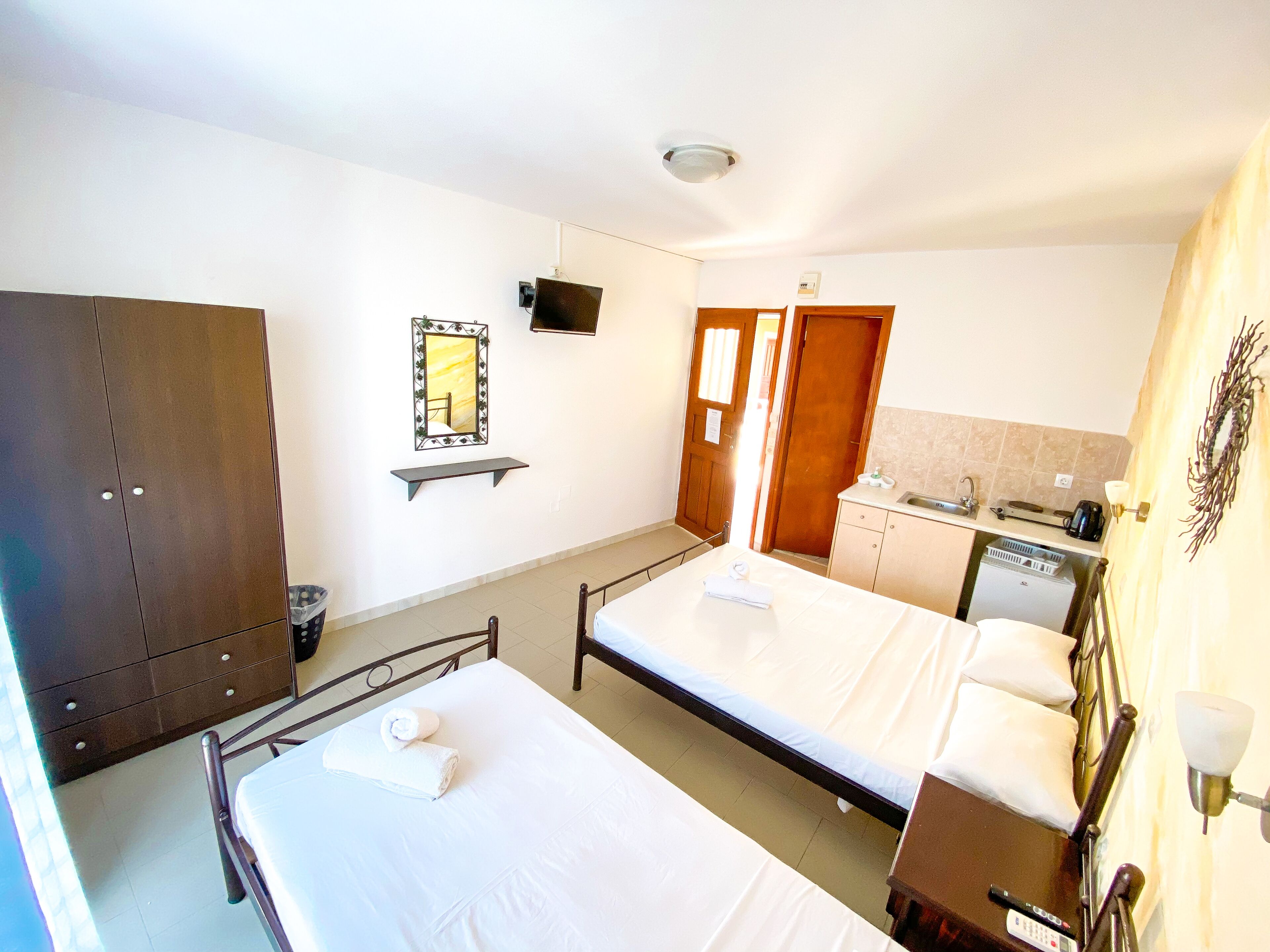 A hotel room with two beds, a wardrobe, a mirror, a television, a bathroom with a sink and a kettle, and a small shelf.