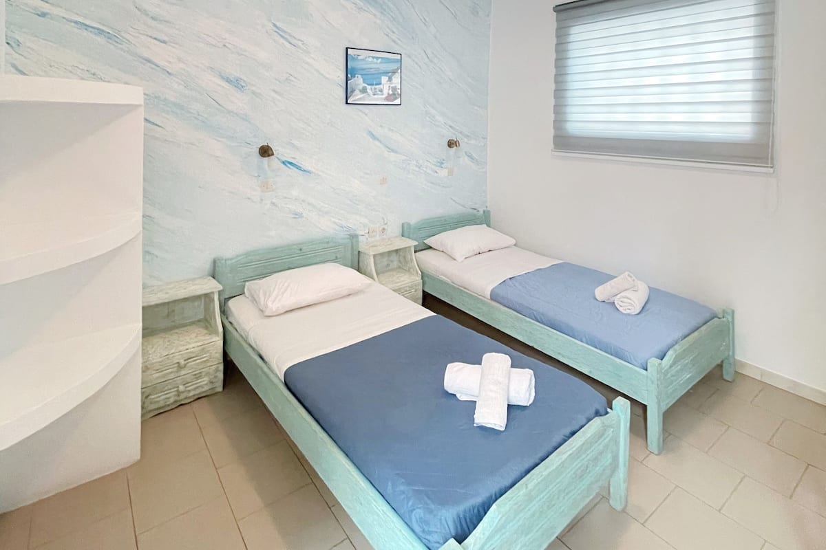Une chambre avec deux lits, une fenêtre avec des stores, un tableau accroché au mur et un luminaire au plafond.