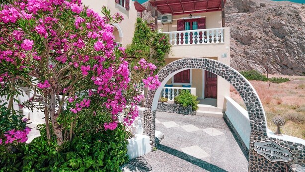 A building with a stone archway entrance, a balcony with red railings, and pink flowers in the foreground. - Villa Dima (Santorini)