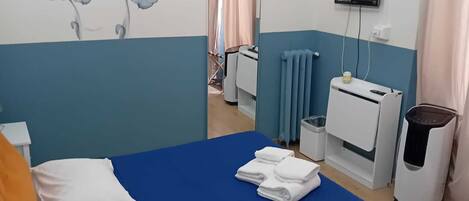 Un dormitorio con una colcha azul, almohadas blancas y una almohada naranja. Hay un televisor en la pared, un radiador y un cubo de basura.